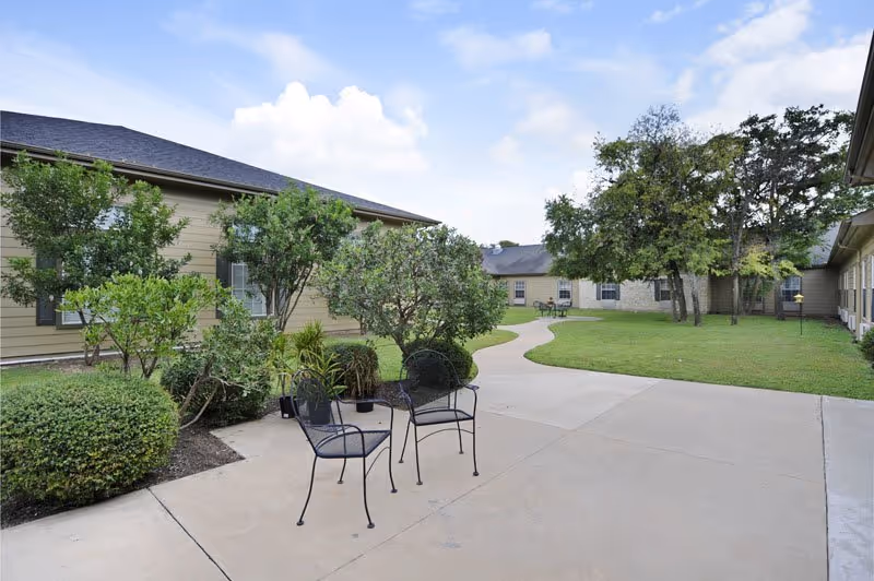Outdoor courtyard with two metal chairs on a paved patio, a winding walkway, lawn, trees, and single-story buildings surrounding the area.
