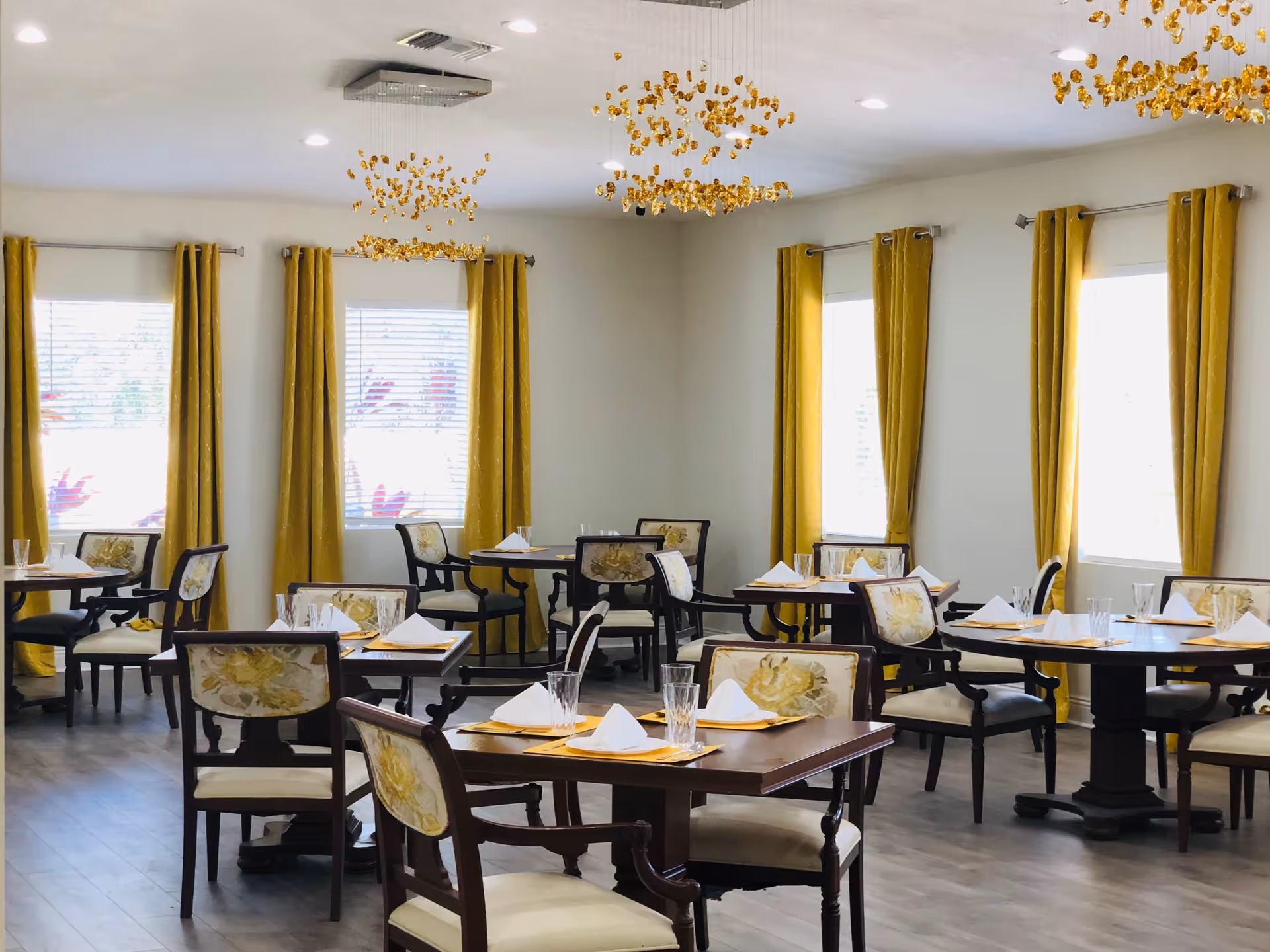 A bright dining room with multiple wooden tables and chairs. Each table is set with yellow placemats, white folded napkins, and clear drinking glasses. The chairs have floral upholstery on the backrests. The room has large windows with yellow curtains and modern decorative ceiling lights.