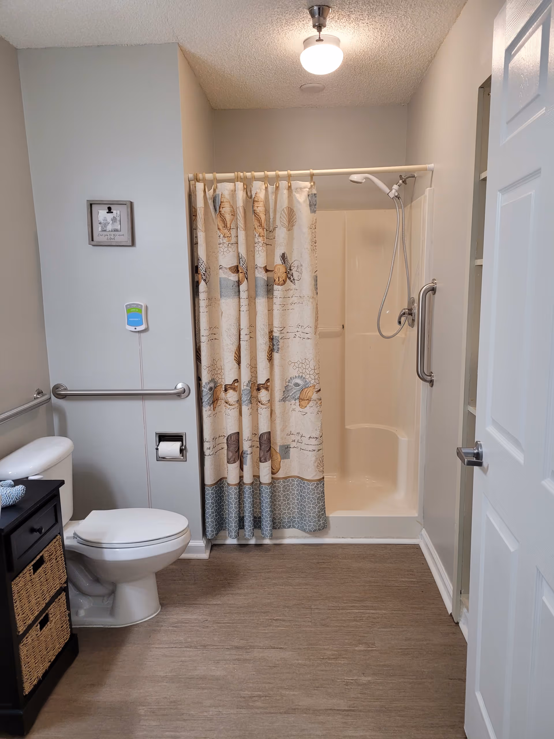 A clean, accessible bathroom featuring a white toilet with grab bars on the wall beside it, a shower with a patterned curtain, and a small black cabinet with wicker drawers. The floor is wood-style vinyl, and the walls are painted light gray. A ceiling light fixture illuminates the space.