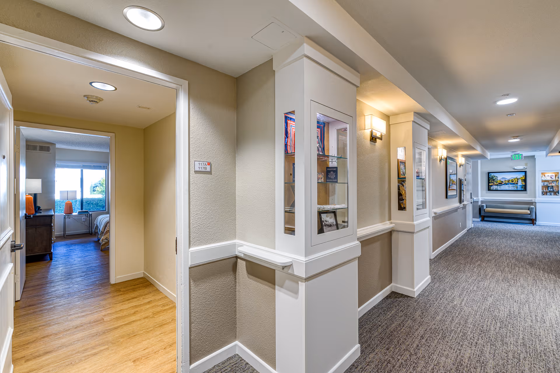 Interior hallway of a senior living facility with beige walls and carpeted floor. On the left, an open door reveals a bedroom with wooden flooring, a bed, a dresser, and a window with a view of greenery outside. The hallway features display cases built into the walls, wall-mounted lights, framed pictures, and a bench at the far end.
