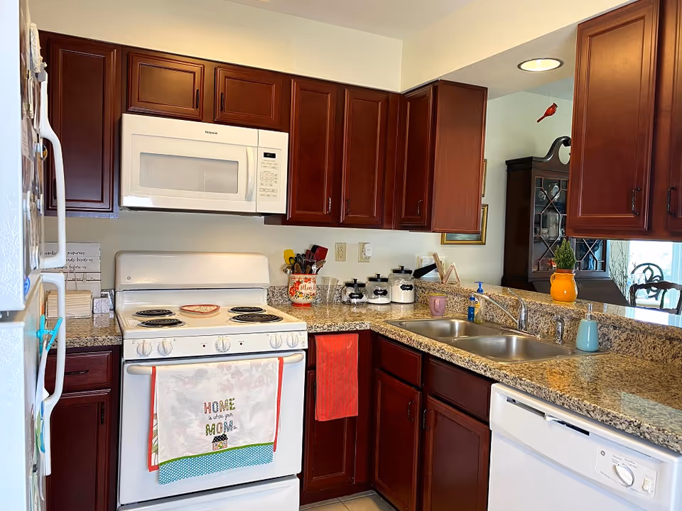 Well-lit kitchen with dark wood cabinets, a white stove and microwave, granite countertops, a double sink, and small decorative items.