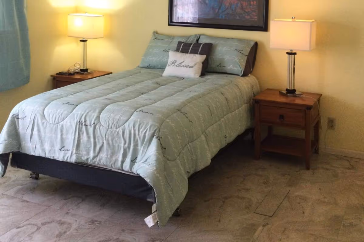 A bedroom with a neatly made bed featuring light blue bedding with script writing. There are two wooden nightstands on either side of the bed, each with a modern table lamp. The walls are painted light yellow, and a framed picture hangs above the bed.