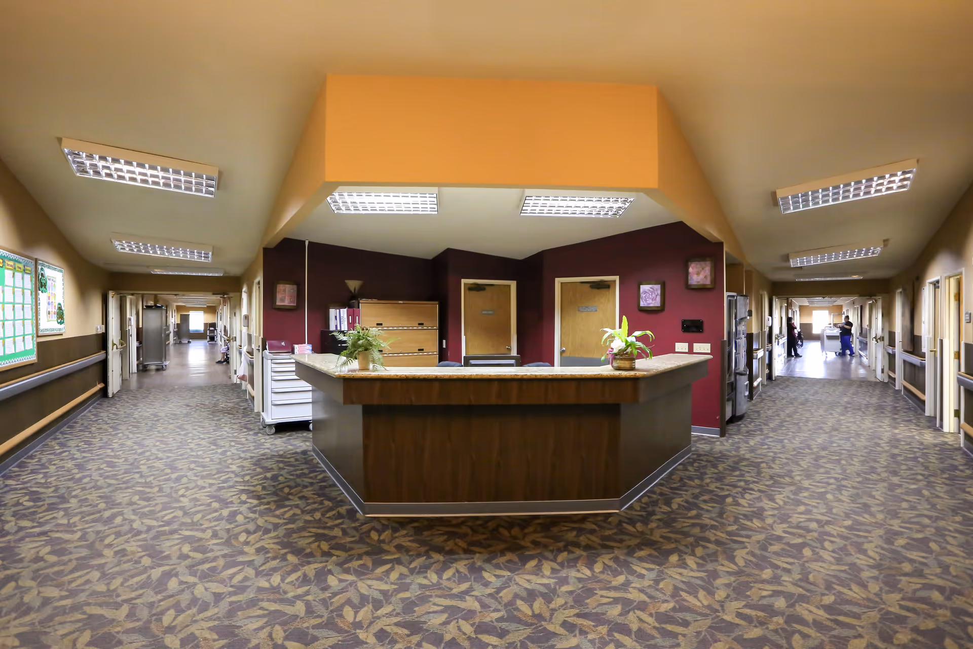 Interior view of a senior living facility hallway with a central nurses' station featuring a wooden counter and two potted plants. The hallway has patterned carpet flooring, beige walls with handrails, and multiple doors along both sides. Fluorescent ceiling lights illuminate the area, and a few staff members are visible in the distance.