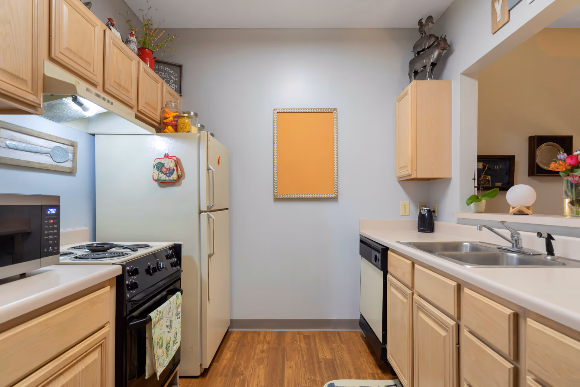 A compact kitchen with light wood cabinets, a white refrigerator, a black stove with an oven, a microwave, and a double sink. The kitchen has light-colored countertops and wood flooring. There is a small bulletin board on the wall and some decorative items on top of the cabinets and counter.