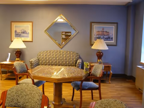 A cozy sitting area with a patterned loveseat against a gray wall, flanked by two wooden side tables each holding a lamp with a beige shade. In front of the loveseat is a wooden octagonal table surrounded by four upholstered chairs with patterned fabric. Above the loveseat is a diamond-shaped mirror, and on either side of the mirror are framed pictures. The floor is wooden, and there is a window on the right side letting in natural light.