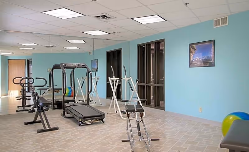 A fitness room with exercise equipment including a treadmill, stationary bike, and other workout machines. The room has light blue walls, a tiled floor, large windows, and a wall mirror reflecting the equipment. A colorful exercise ball is partially visible on the right side.