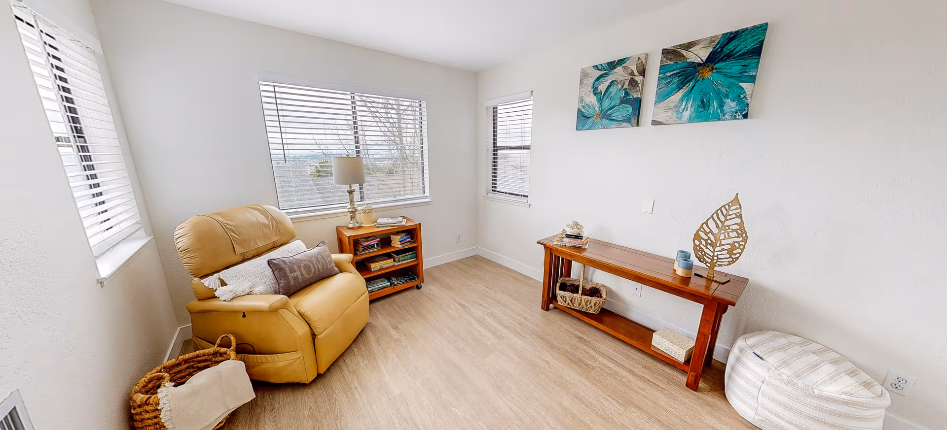 A bright, cozy living room with light wood flooring and white walls. The room features a tan recliner chair with pillows, a small wooden bookshelf with a lamp and books, a long wooden console table with decorative items, a woven basket, a striped pouf, and two large floral paintings on the wall. There are three windows with white blinds letting in natural light.
