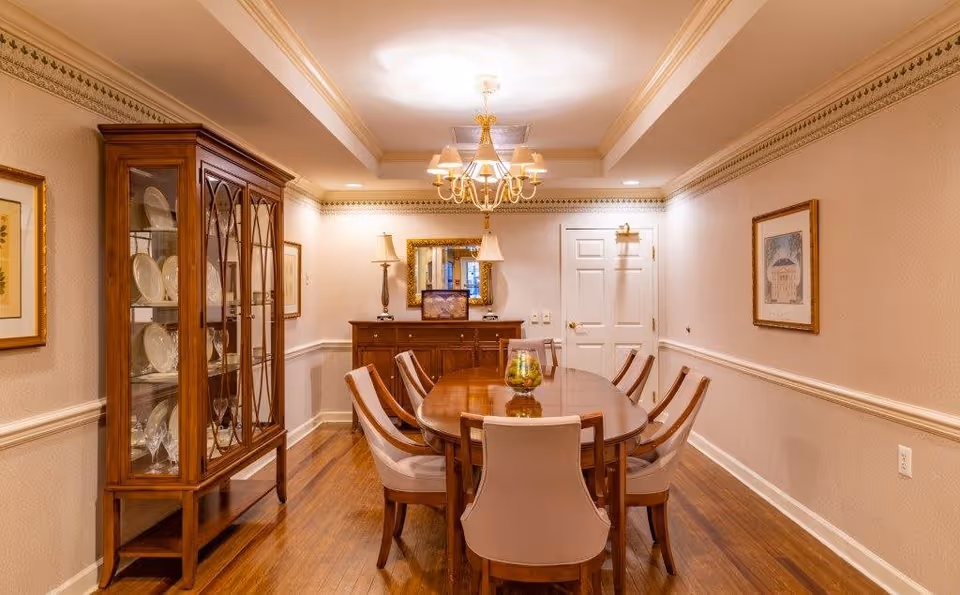 A formal dining room with a wooden dining table surrounded by eight upholstered chairs. A glass-front china cabinet filled with plates and glassware is on the left side. The room has wooden flooring, beige walls with decorative trim, framed artwork, a chandelier hanging from the ceiling, and a sideboard with two lamps and a mirror above it at the far end.