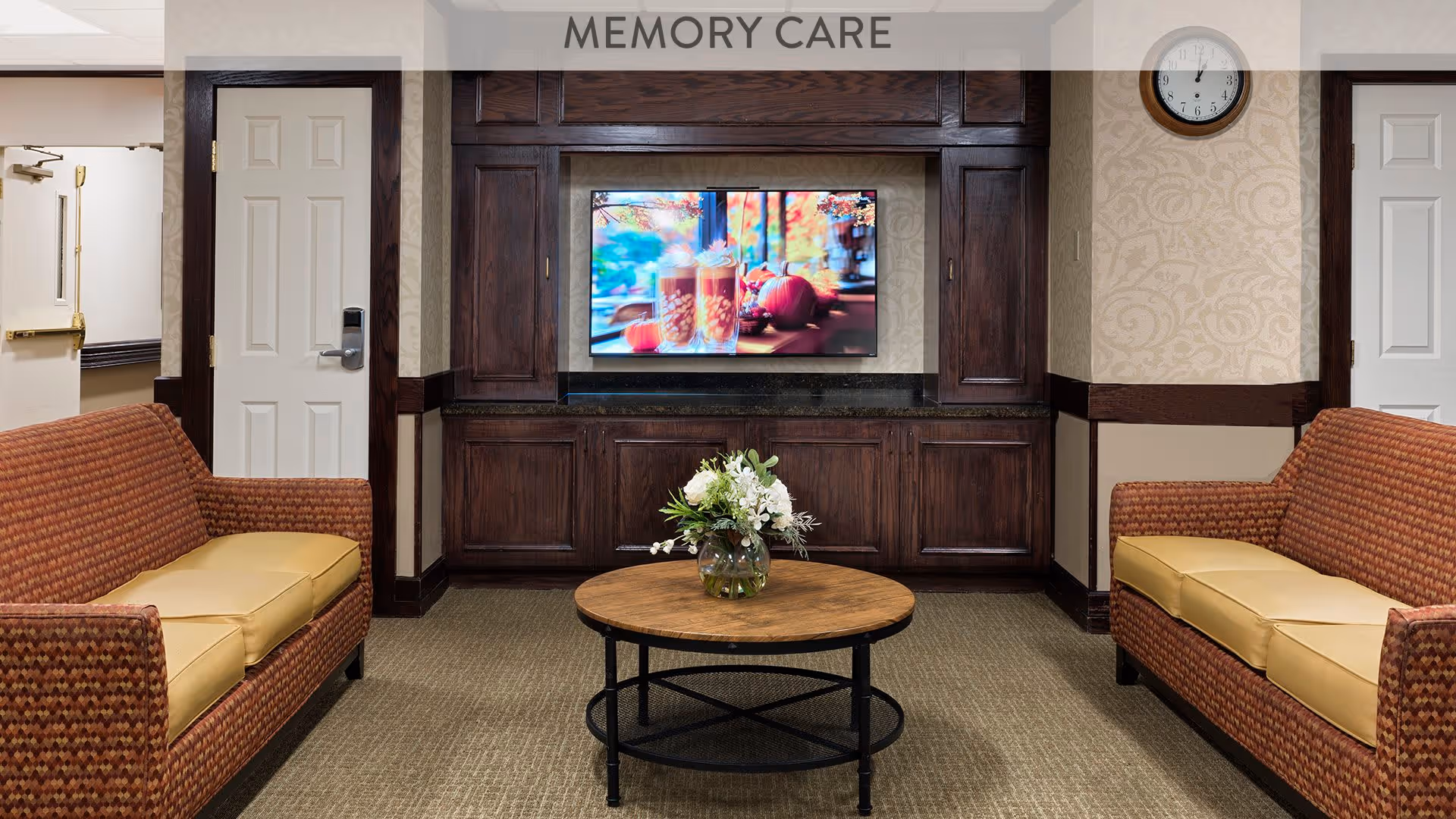 A cozy memory care lounge area with two patterned sofas facing each other, a round wooden coffee table with a glass vase of white flowers in the center, a wall-mounted flat screen TV displaying a fall-themed image, dark wood cabinetry, beige patterned wallpaper, and a clock on the wall showing 12:00.