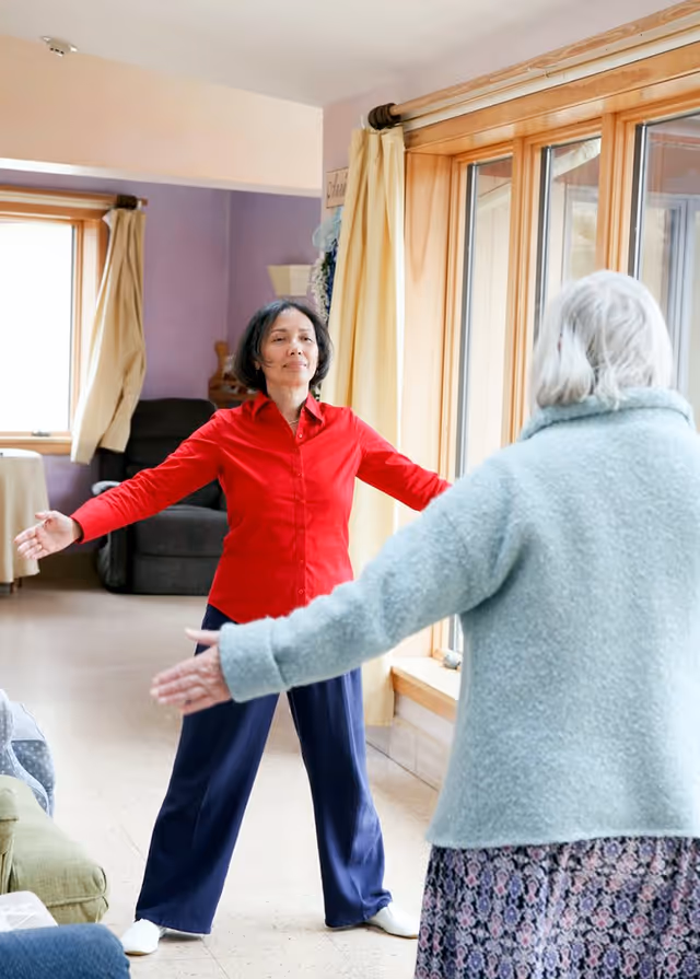 Two women standing in a living room with large windows and curtains, facing each other with arms outstretched as if exercising or stretching. One woman is wearing a red shirt and dark pants, the other is wearing a light blue sweater and a patterned skirt.