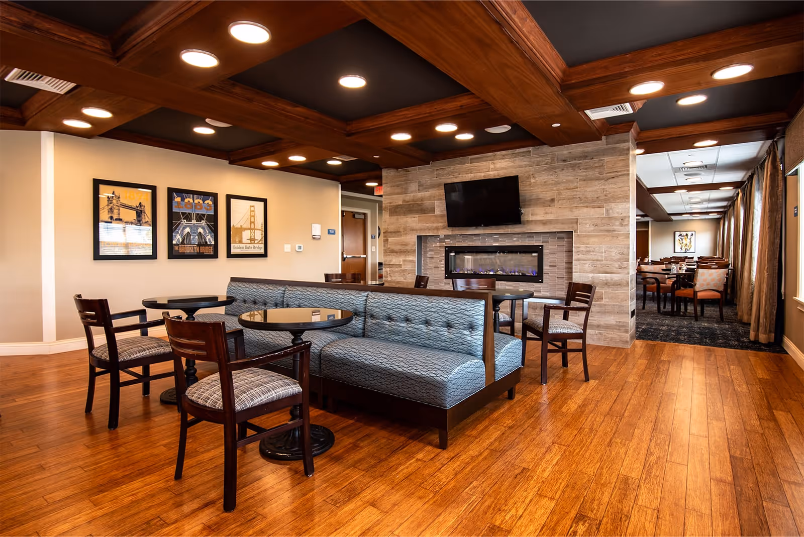 A cozy common area in Woodland Terrace of Carmel featuring a blue cushioned bench seating around circular tables, wooden chairs with patterned cushions, a wall-mounted flat screen TV above a modern electric fireplace with a stone surround, warm wooden flooring, and framed pictures on the wall. The ceiling has wooden beams with recessed lighting, and there is an adjoining dining area visible in the background.
