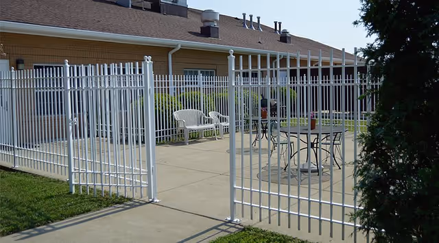 Gated outdoor patio with white metal fencing, plastic and metal chairs and tables beside a brick assisted living building.