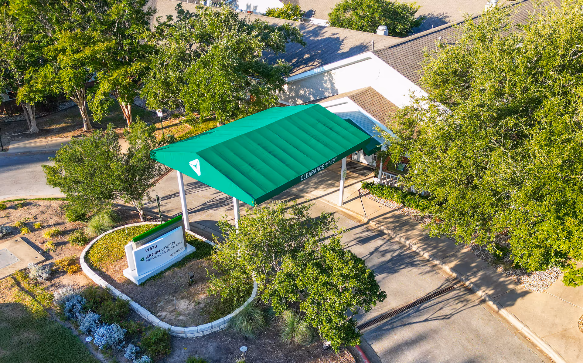 Aerial view of the entrance to Arden Courts - ProMedica Memory Care Community in Austin, featuring a green canopy with clearance height marked, surrounded by trees and landscaping.