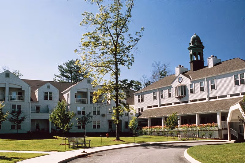 Front exterior of a white multi-story senior living building with a central cupola, lawn, benches, trees, and a curved driveway.
