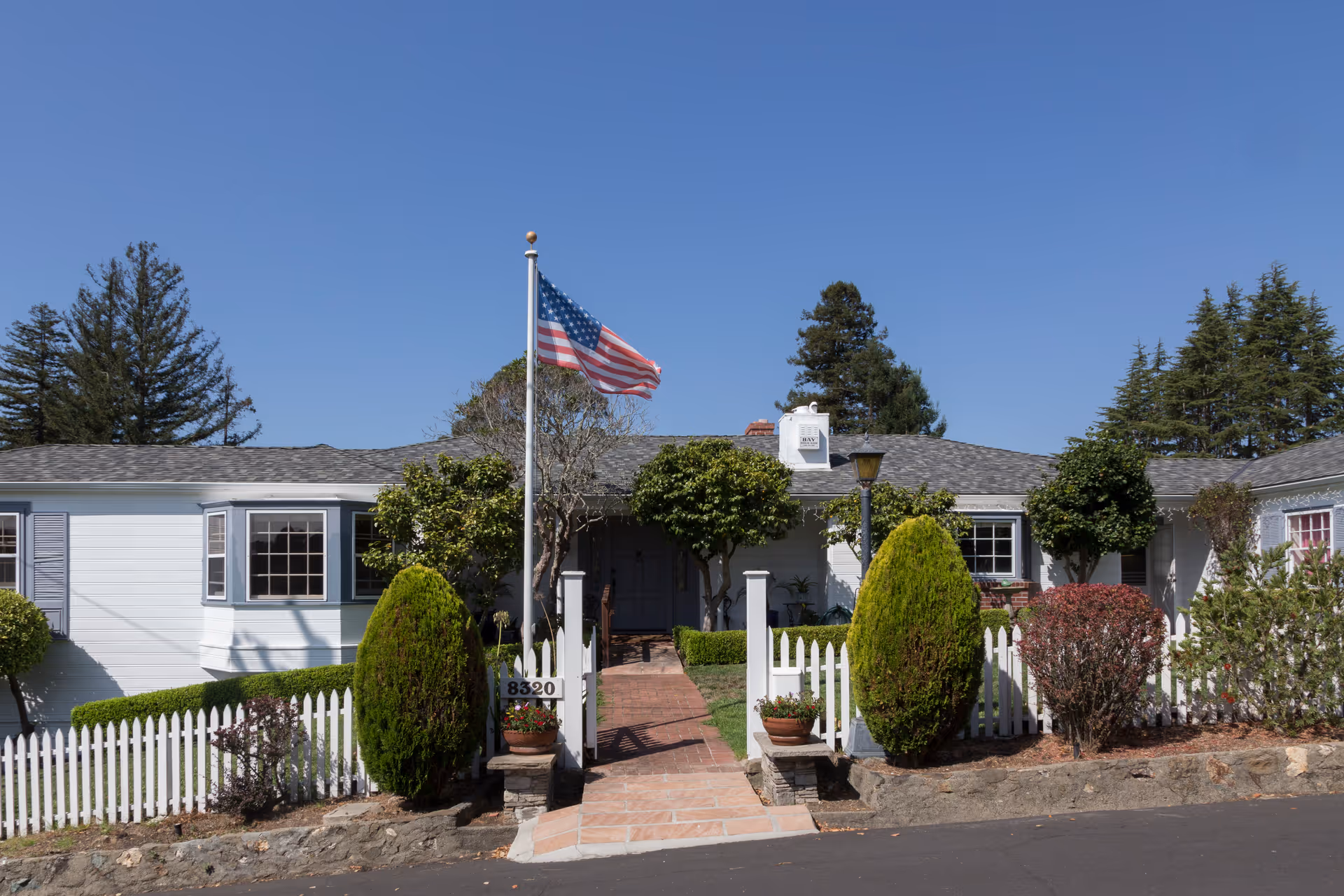 Front exterior view of a single-story assisted living home with a white picket fence, neatly trimmed bushes, an American flag on a flagpole, and a clear blue sky.