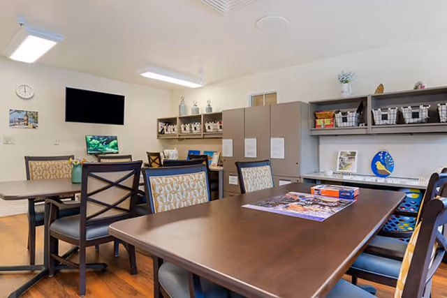 Communal activity room with wooden tables and chairs, storage shelves, a wall-mounted TV, and a puzzle on the table.