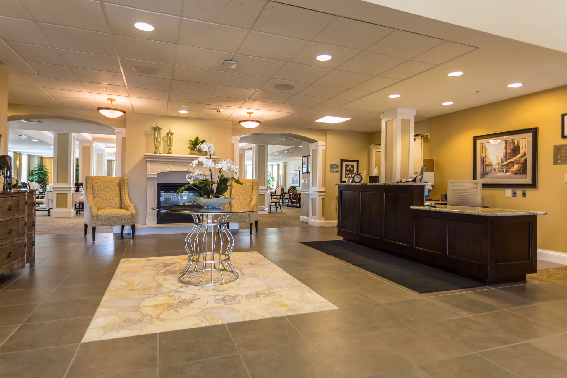 A spacious senior living facility lobby with a dark wooden reception desk on the right, a decorative table with a flower arrangement in the center on a floral rug, two upholstered armchairs near a fireplace, and warm yellow walls with framed artwork.
