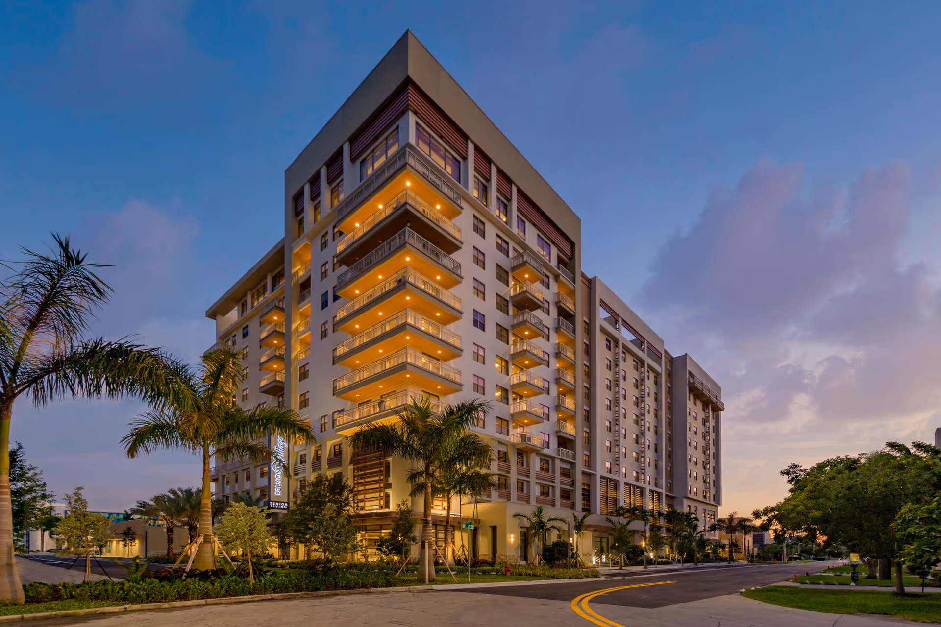 Exterior view of a modern multi-story senior living facility building at dusk with balconies, palm trees, and illuminated lights along the facade and entrance area.