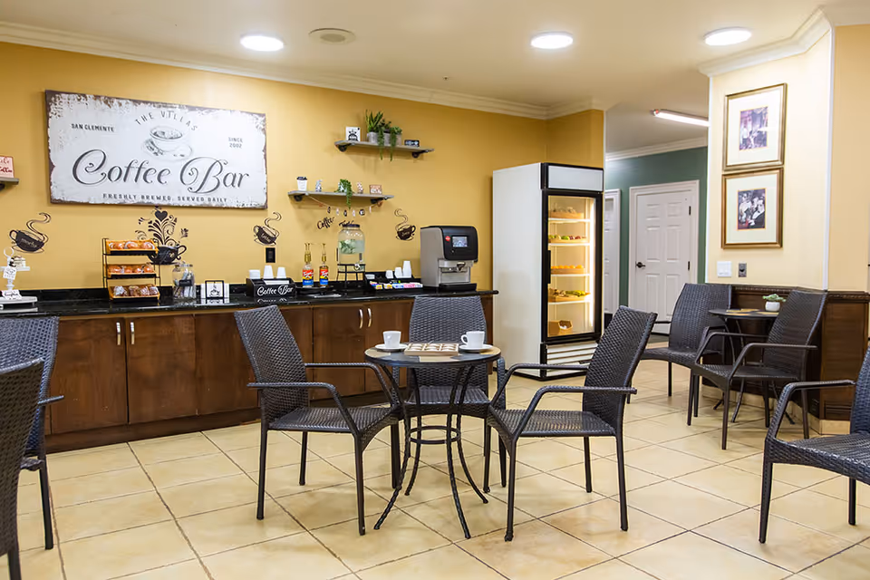 A cozy coffee bar area in San Clemente Villas with a sign reading 'Coffee Bar' on the wall. The space features a counter with coffee supplies, a water dispenser, and a refrigerated display case with snacks. There are several wicker chairs and small round tables with coffee cups on them, set on a tiled floor with warm yellow walls and framed pictures.