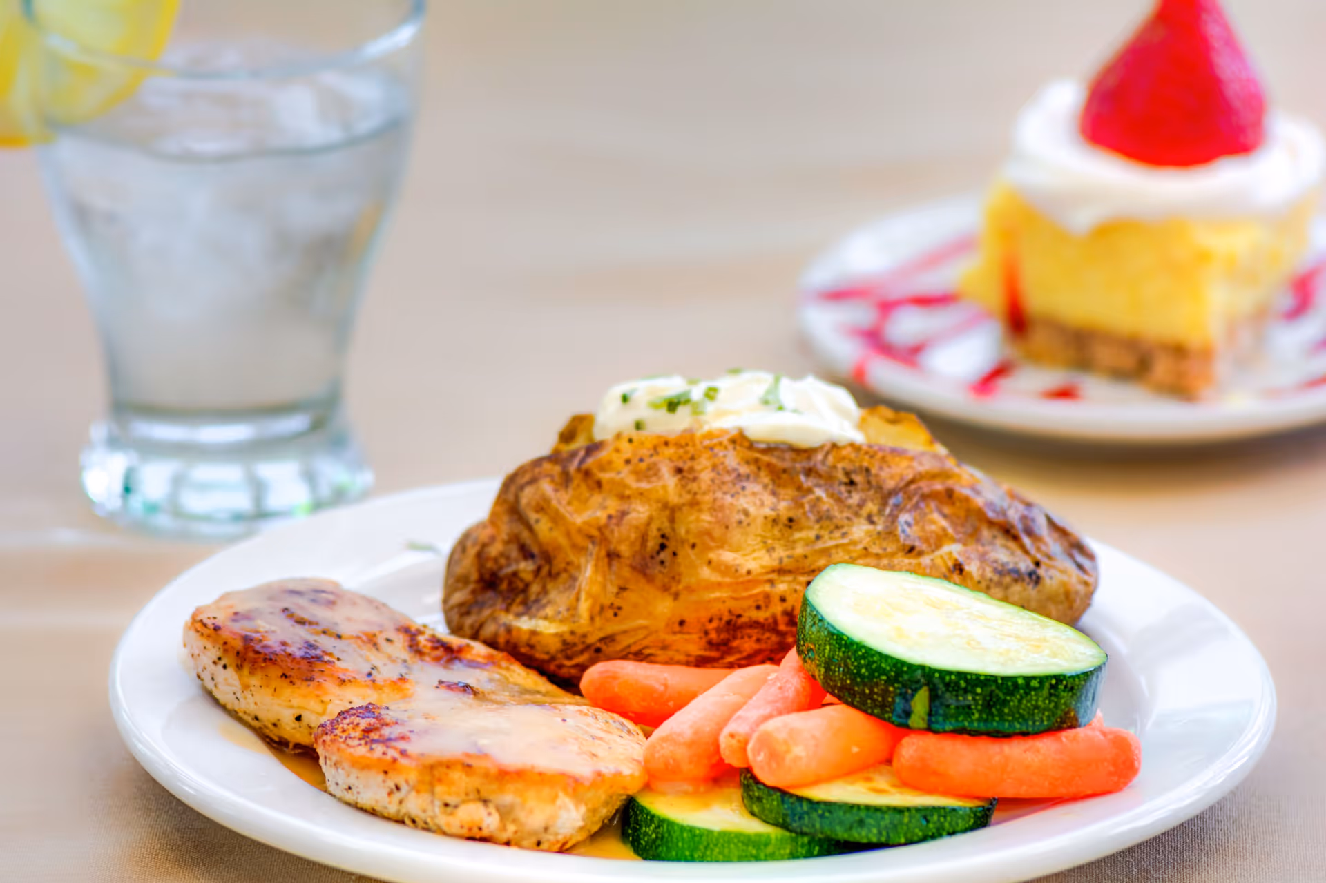 A plated meal with a baked potato topped with sour cream, grilled chicken, sliced zucchini and baby carrots, with a glass of water and a dessert in the background.