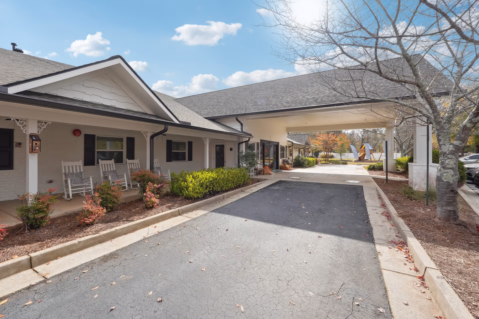 Exterior view of a senior living facility with a covered driveway entrance. The building has white brick walls, a gray shingled roof, and a porch with white rocking chairs. There are small bushes and trees around the driveway, and a playground is visible in the background under a partly cloudy sky.
