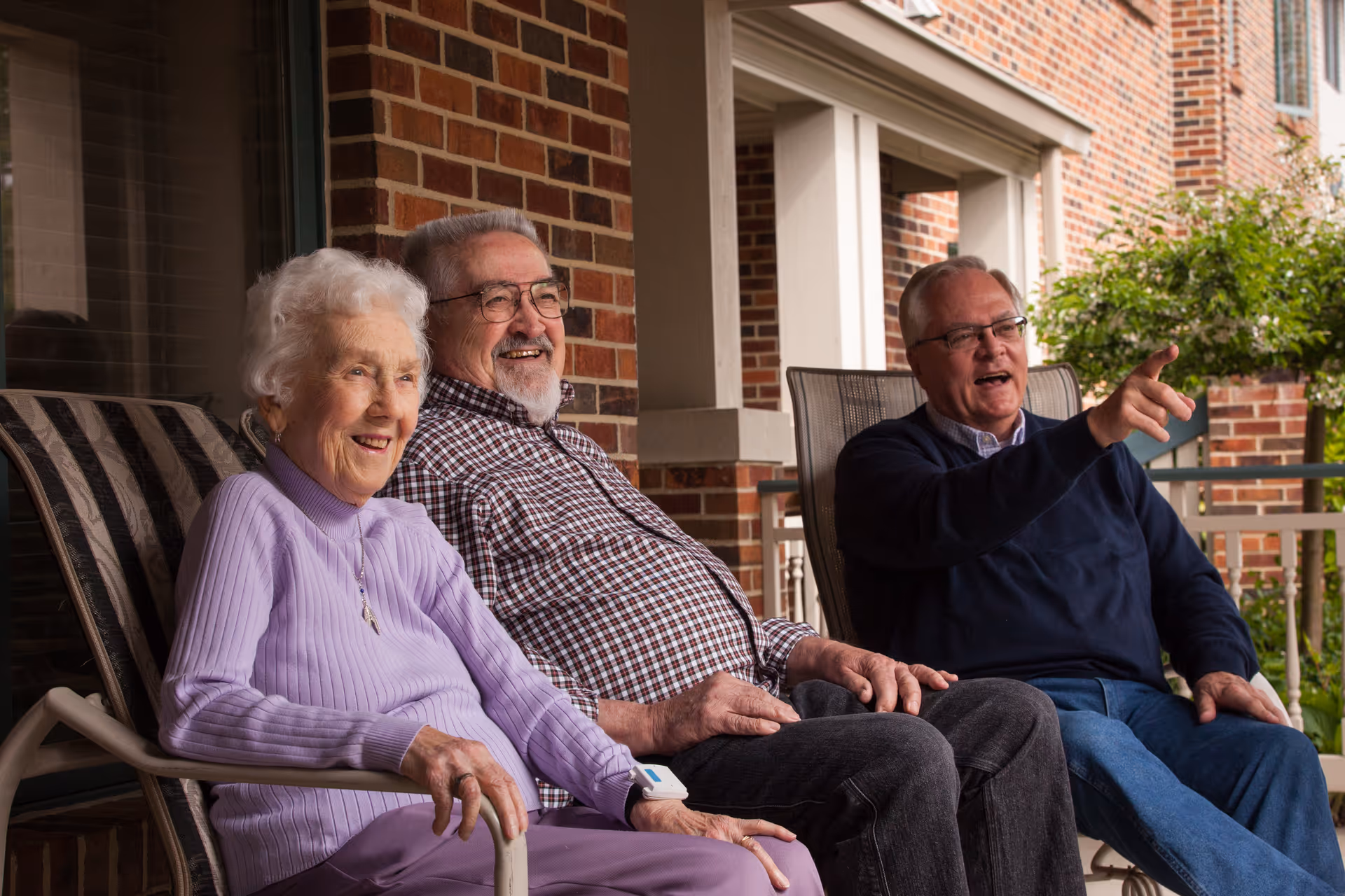 Three elderly people sitting on chairs on a porch outside a brick building. Two men and one woman are smiling and engaging in conversation, with one man pointing at something in the distance. There is greenery visible in the background.