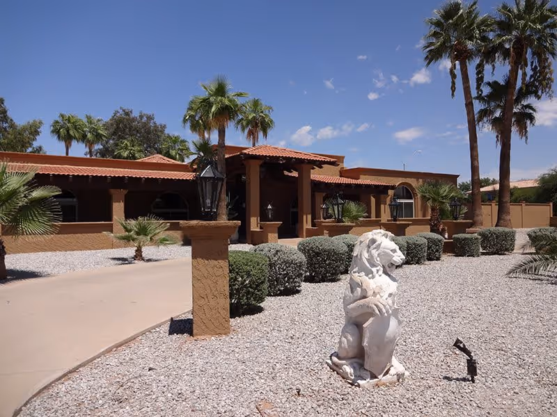 Exterior view of The Sanctuary of Scottsdale facility featuring a desert-style landscape with gravel ground cover, trimmed bushes, palm trees, and a white lion statue in the foreground. The building has a brown stucco finish with a red tile roof and arched windows under a clear blue sky.