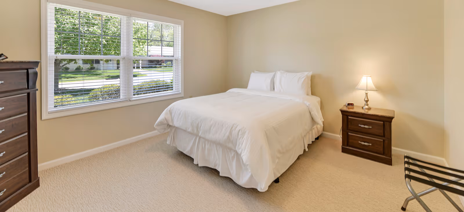 A bedroom with a neatly made bed with white bedding, a wooden nightstand with a lamp and clock, a wooden dresser, a large window with white blinds showing greenery outside, and beige walls and carpet.