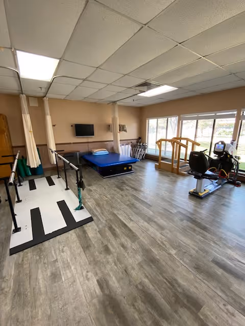 Bright therapy room with parallel bars, a blue mat, practice stairs and an exercise bike beside large windows.