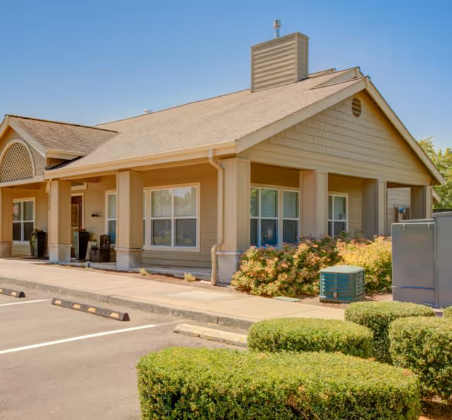 Front exterior view of a single-story senior living facility with tan siding and shrubs.