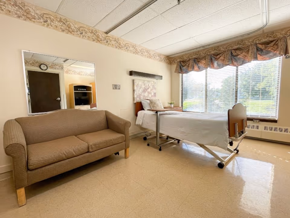 A bright room with a hospital-style bed next to a large window with blinds and a floral valance. There is a beige couch on the left side and a large mirror on the wall reflecting a door and a TV cabinet. The floor is tiled and the walls are light-colored with a decorative border near the ceiling.