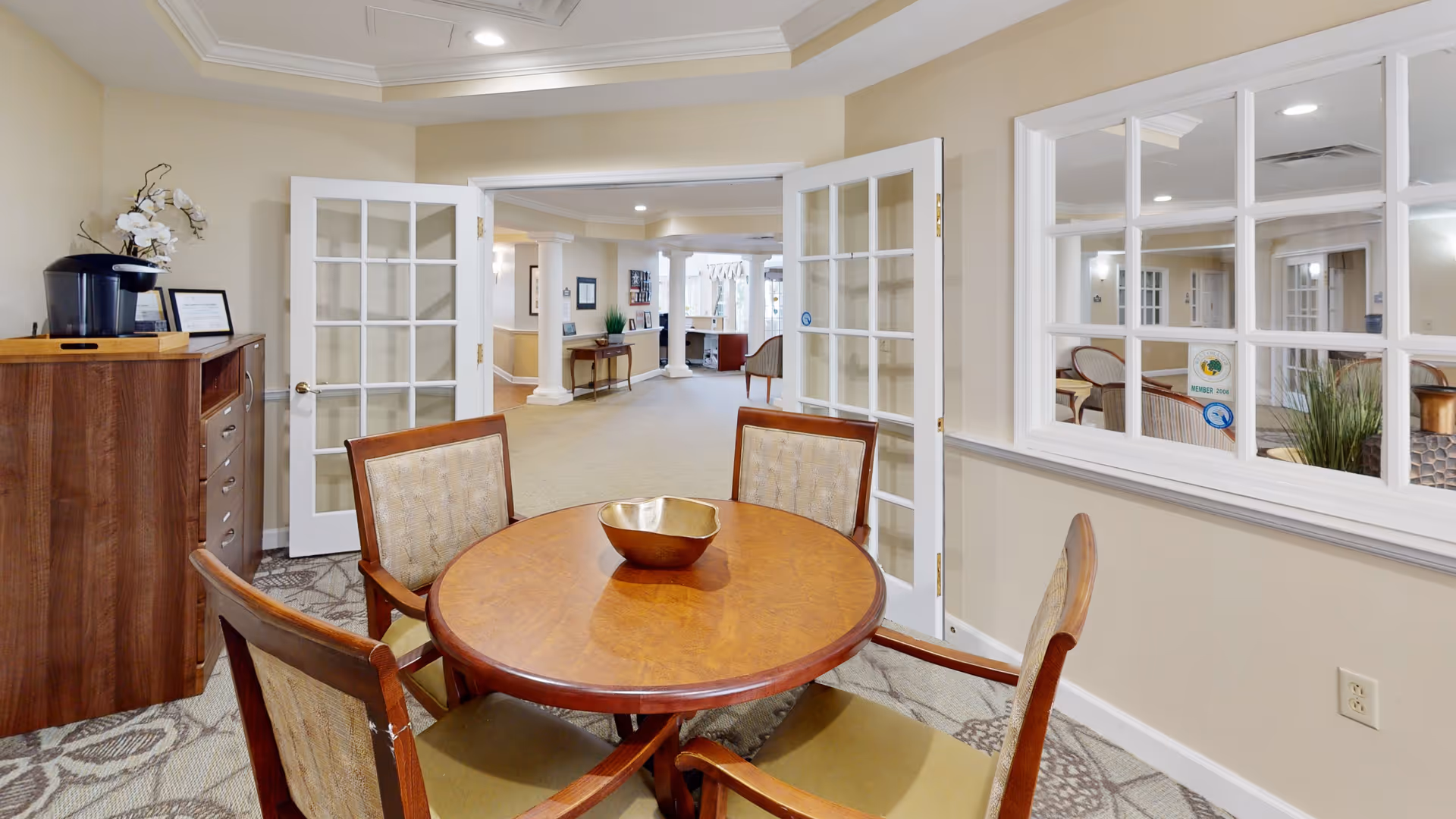 A small round wooden table with four cushioned chairs around it in a well-lit room. There is a decorative bowl on the table. Behind the table, there is a wooden cabinet with a coffee maker and a framed certificate or picture. The room has beige walls, carpeted floor, and white trim. French doors open to a hallway with columns and additional seating visible in the background.