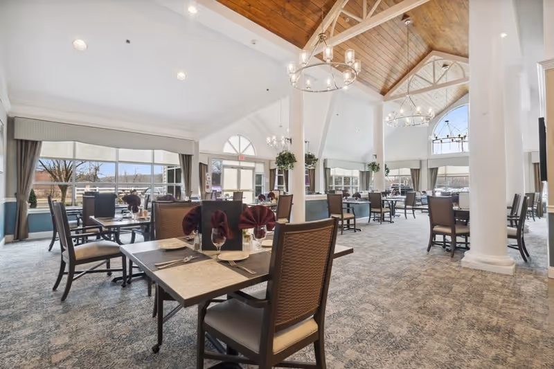 A spacious senior living dining room with high vaulted wooden ceilings, large windows letting in natural light, multiple dining tables set with plates, glasses, and folded napkins, and elegant chandeliers hanging from the ceiling.