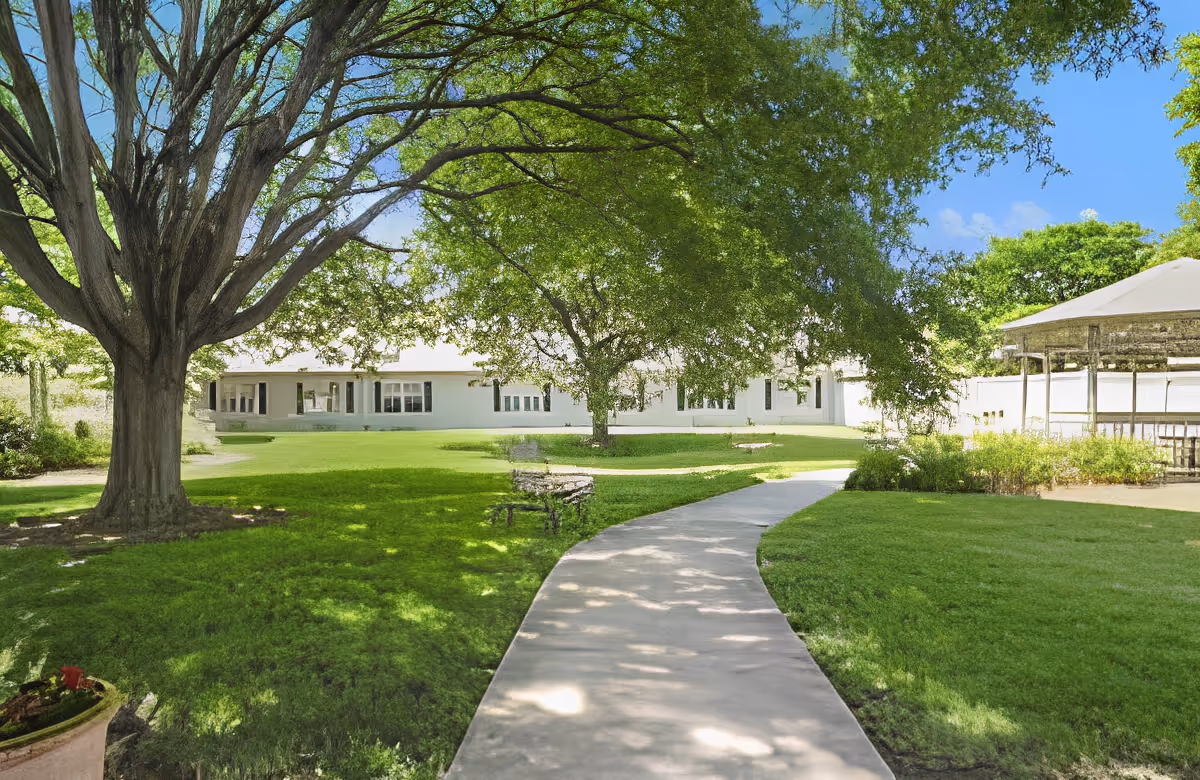 A paved walkway curves through a green lawn with large leafy trees providing shade. In the background, there is a white building with multiple windows and a covered outdoor seating area. The sky is clear and blue.
