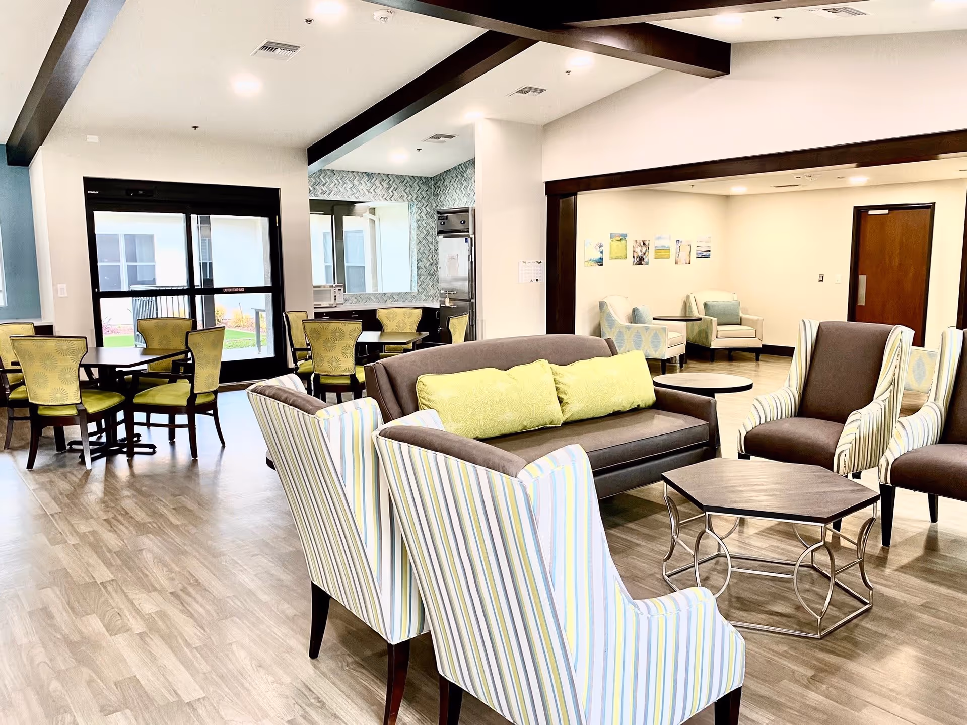 A bright and spacious common area in a senior living facility featuring a seating arrangement with a brown sofa with green cushions, striped armchairs, and a hexagonal coffee table. In the background, there are several dining tables with green cushioned chairs near large windows and a glass door leading outside. The walls are decorated with small framed pictures, and the flooring is wood-style laminate.