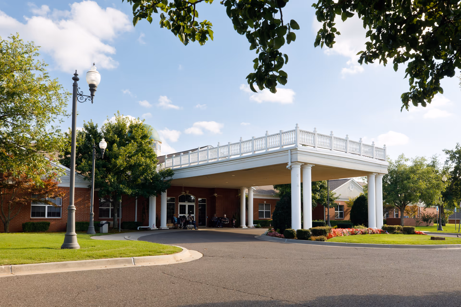 Exterior view of Arbor House of Midwest City showing a covered entrance with white columns, a circular driveway, green trees, and a well-maintained lawn under a partly cloudy sky.