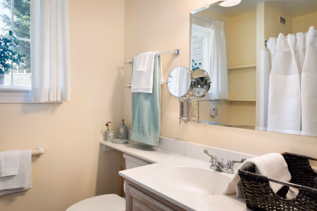 A clean and bright bathroom with a white sink and countertop, a large mirror, a small round magnifying mirror attached to the wall, a towel rack with white and light blue towels, a window with white curtains, and a black basket holding white towels on the counter.