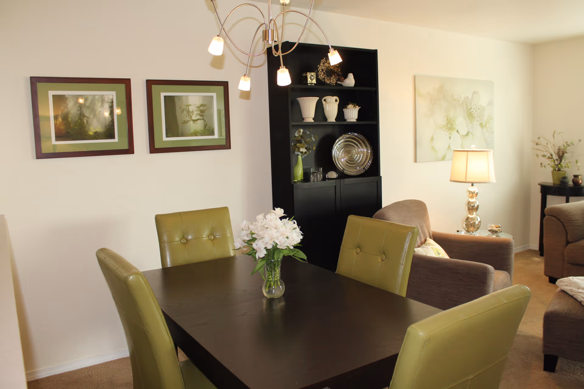 A cozy interior living and dining area featuring a dark wooden dining table with four green cushioned chairs. A vase with white flowers is placed on the table. In the background, there is a black shelving unit with decorative items, a beige armchair, a table lamp with a glass base, and framed artwork on the walls.