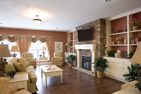 A cozy living room with beige sofas and armchairs arranged around a white coffee table on a wooden floor. The room features a stone fireplace with a mounted flat-screen TV above it, built-in white shelves with decorative items and plants, and large windows with patterned valances allowing natural light to fill the space.