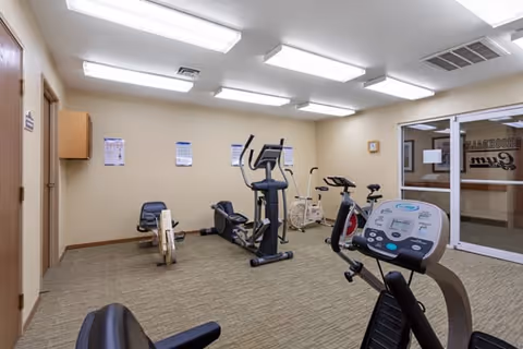 A small exercise room with various fitness equipment including stationary bikes, an elliptical machine, and a rowing machine. The room has beige walls, carpeted floor, fluorescent ceiling lights, and a glass door leading to another area labeled 'Brookdale Gym'.