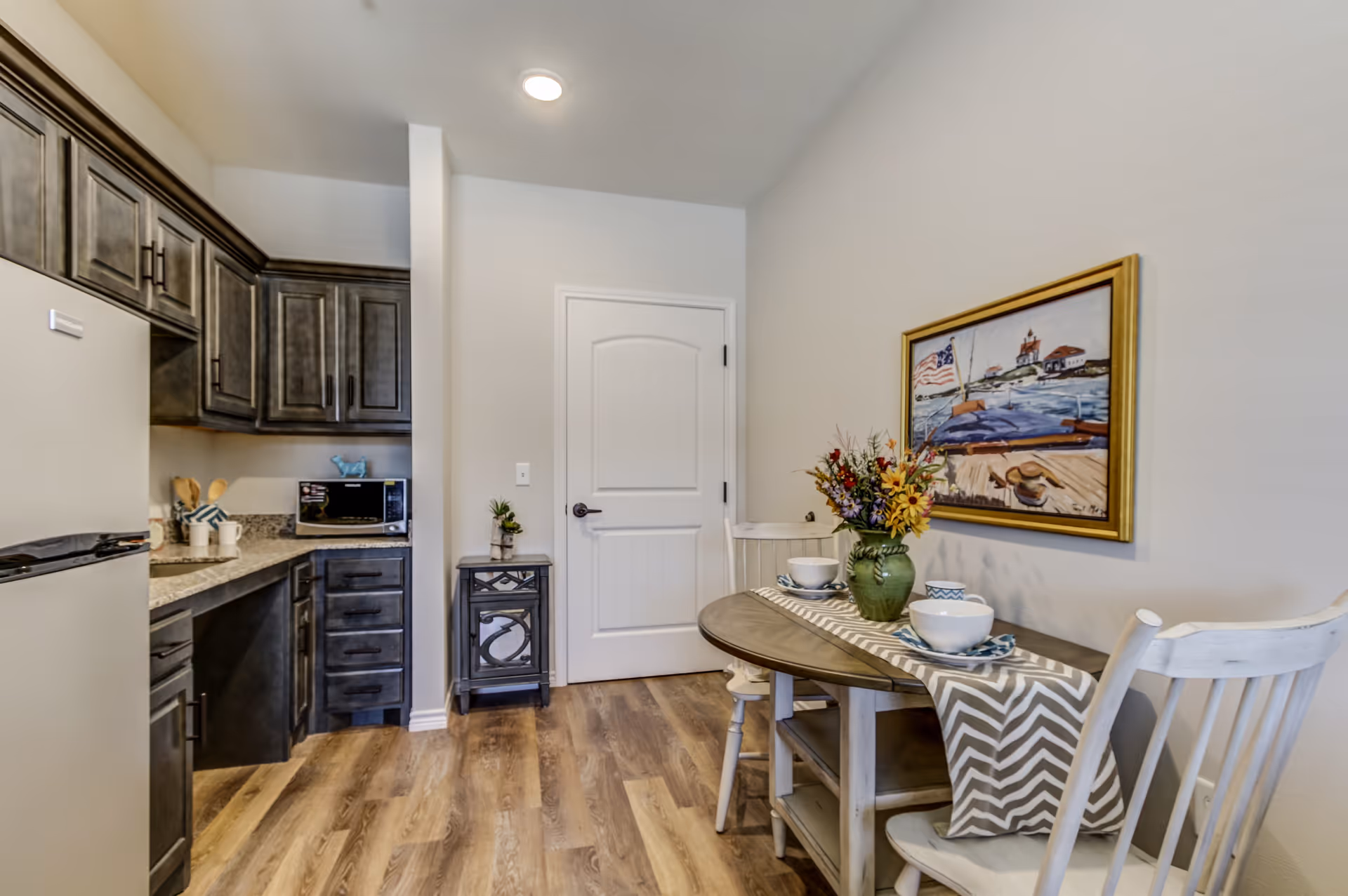 Kitchenette with dark cabinets, a small round dining table set for two and a framed painting on the wall.