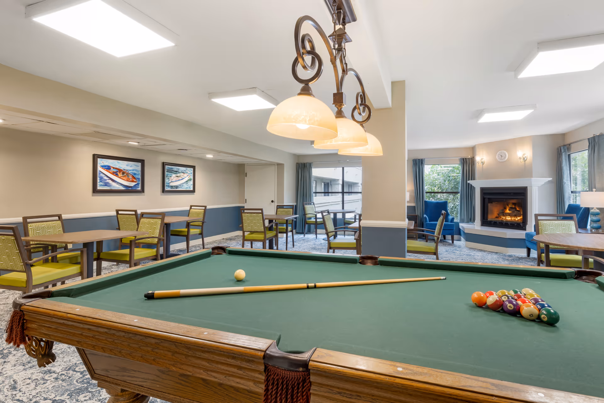 A senior living facility common area featuring a pool table with a cue stick and billiard balls arranged for a game. The room has multiple tables and chairs with green cushions, a fireplace with a fire burning, blue armchairs, large windows with blue curtains, and framed pictures of boats on the wall.