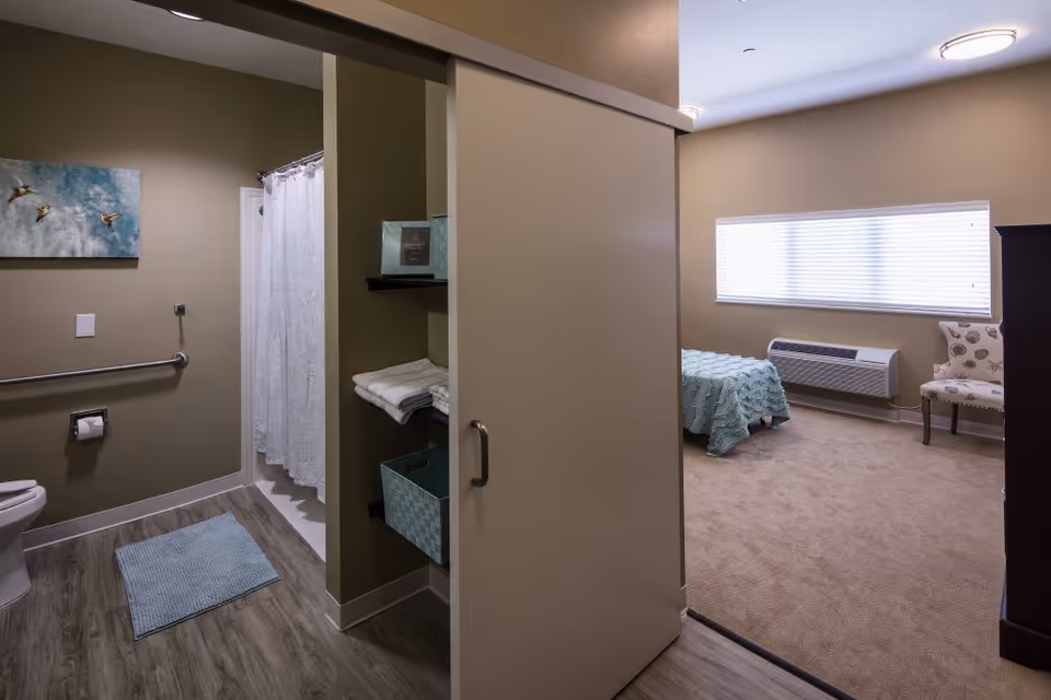 View of a memory care facility room showing a bathroom on the left with a toilet, grab bar, shower with a curtain, and a blue bath mat. To the right is a bedroom area with a bed covered in a light blue quilt, a window with blinds, a patterned chair, and a dark wooden dresser. The bathroom and bedroom are separated by a sliding door.