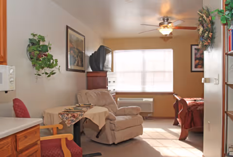 A cozy living space with a recliner chair, a small table covered with a floral tablecloth, a red cushioned chair, a wooden dresser with an old-style TV on top, a large window with blinds, a ceiling fan with light, and part of a bed with a red blanket visible. The room has light-colored walls and carpeted floor, with some framed artwork and decorative plants on the walls.
