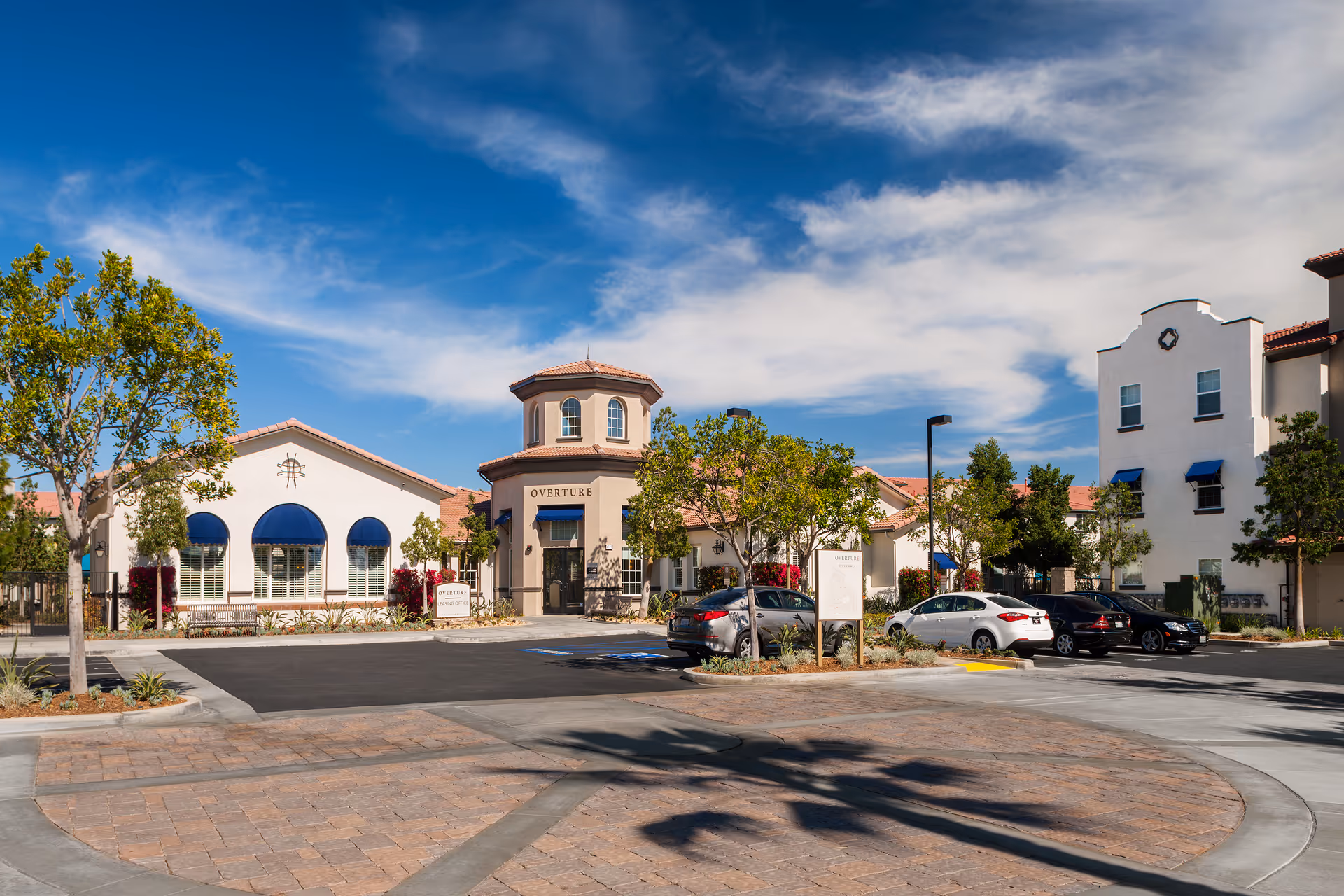 Exterior view of Overture Riverwalk senior living facility on a sunny day with a clear blue sky and some clouds. The building features a Mediterranean architectural style with beige walls, red tile roofs, and blue awnings over the windows. There are several trees and landscaped areas around the parking lot, which has a few parked cars. A sign near the entrance reads 'Overture Leasing Office.'