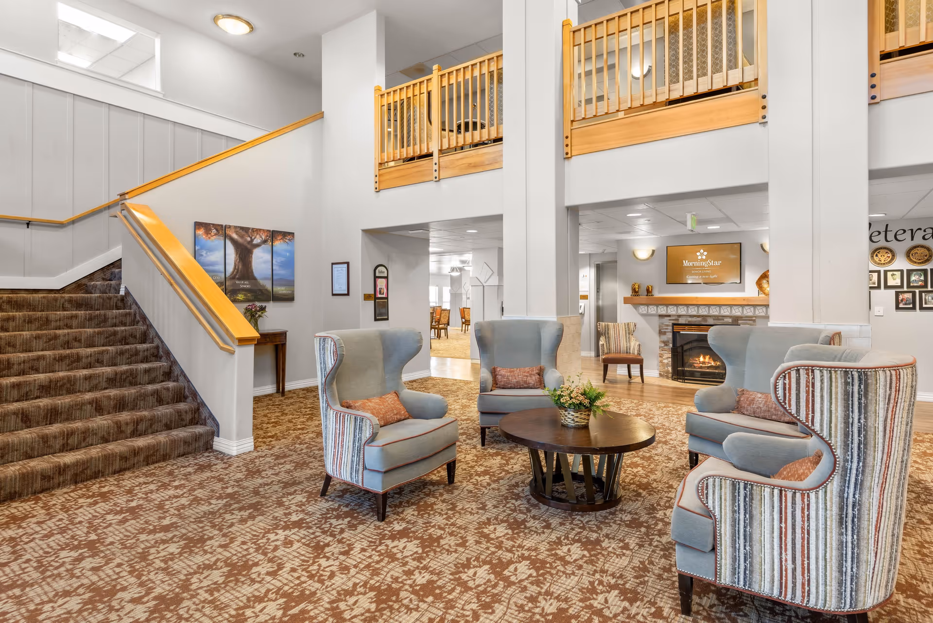 Spacious senior living lobby with armchairs arranged around a coffee table, a fireplace, and a staircase to an upper level.