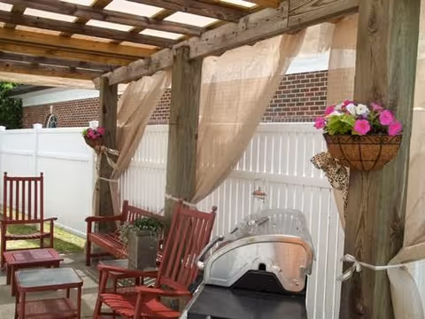 Outdoor patio area with wooden pergola, red wooden chairs and tables, hanging flower baskets with pink and white flowers, beige curtains tied to wooden posts, and a barbecue grill. A white fence and brick building wall are visible in the background.