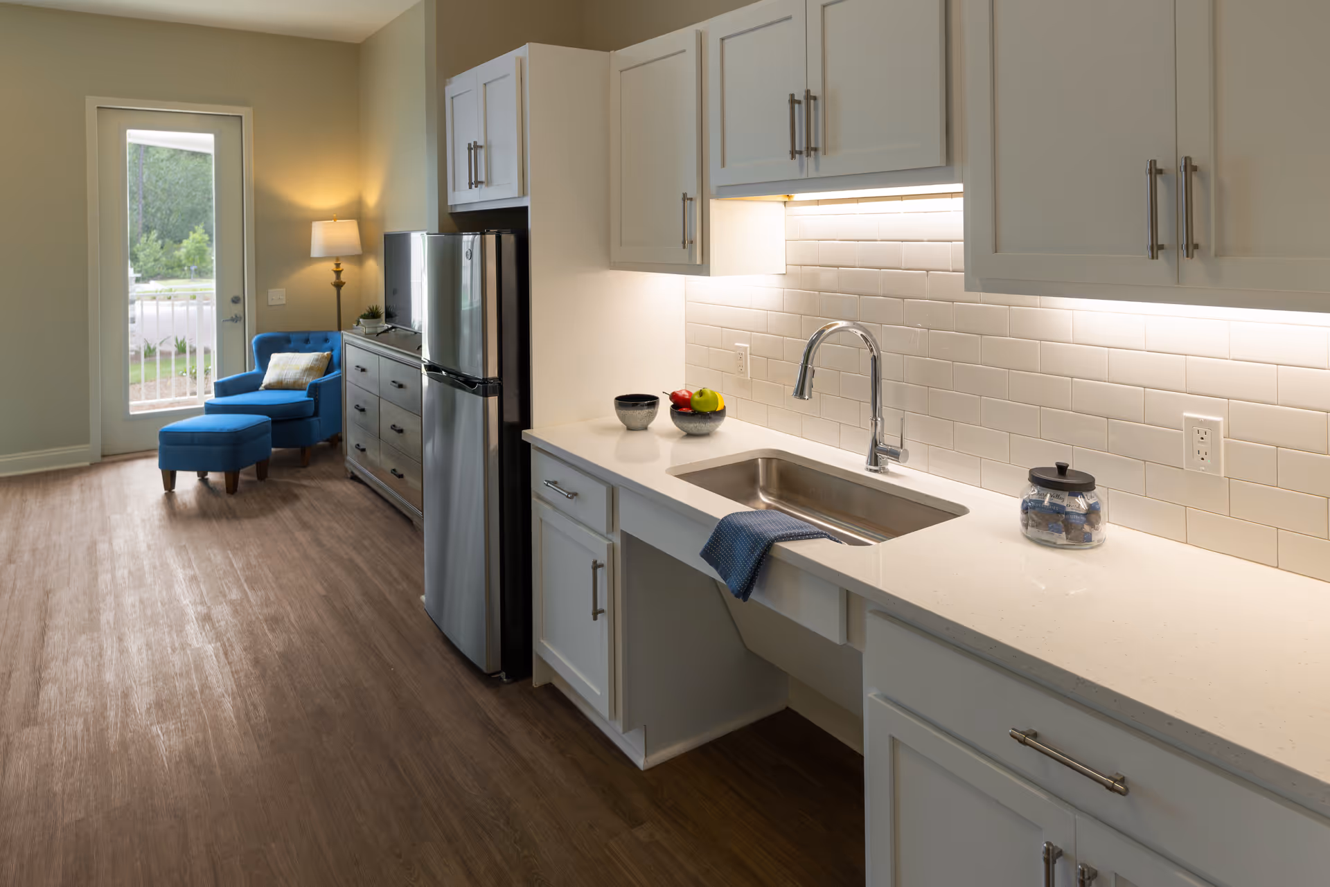 A modern kitchen area with white cabinets, a stainless steel refrigerator, and a sink with a chrome faucet. The countertop is white with a blue towel hanging over the sink. There are bowls with fruit and a jar on the counter. In the background, there is a blue armchair with a matching ottoman and a floor lamp next to a glass door leading outside.