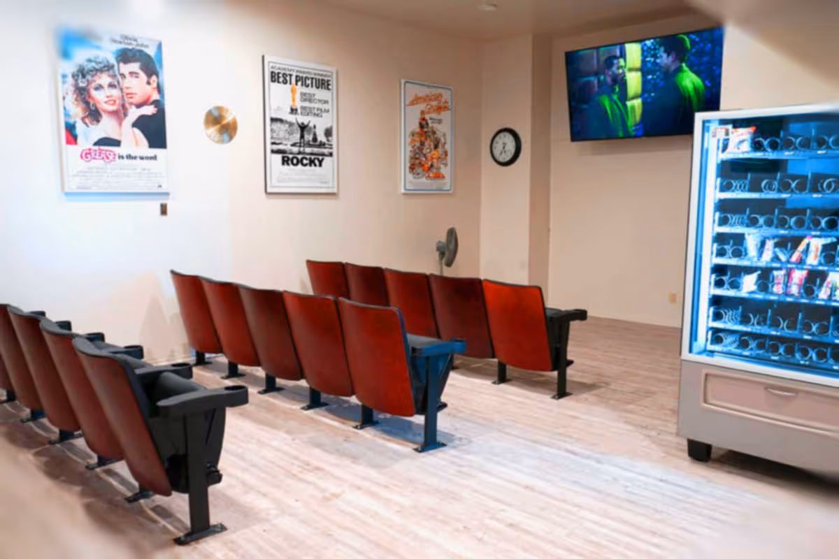 A small movie theater room with two rows of red theater seats facing a wall-mounted TV screen showing a movie scene. On the wall behind the seats are framed posters of classic movies including Grease and Rocky. To the right, there is a vending machine stocked with snacks and drinks. The room has light-colored wooden flooring and a wall clock.