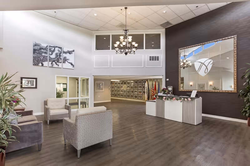 A spacious and well-lit lobby area with modern furniture including armchairs and a small reception desk decorated with flowers. A large mirror with a decorative frame hangs on a dark accent wall behind the desk. The floor is wood, and there is a chandelier hanging from the high ceiling. The back wall features a hallway lined with framed pictures and several flags.